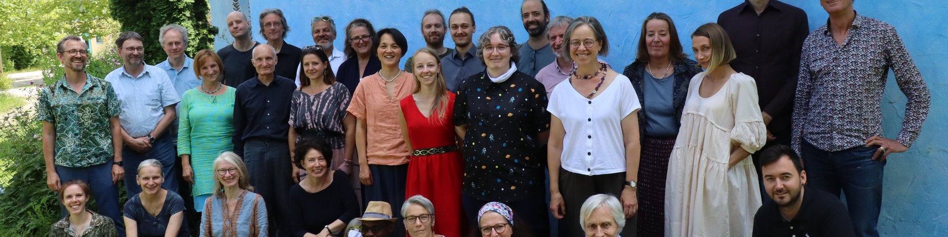 Eine Gruppe von etwa 30 Erwachsenen posiert gemeinsam im Freien vor einer großen blauen Wand. Einige stehen, andere sitzen im Gras und lächeln in die Kamera, während im Hintergrund Bäume und Grünflächen zu sehen sind.