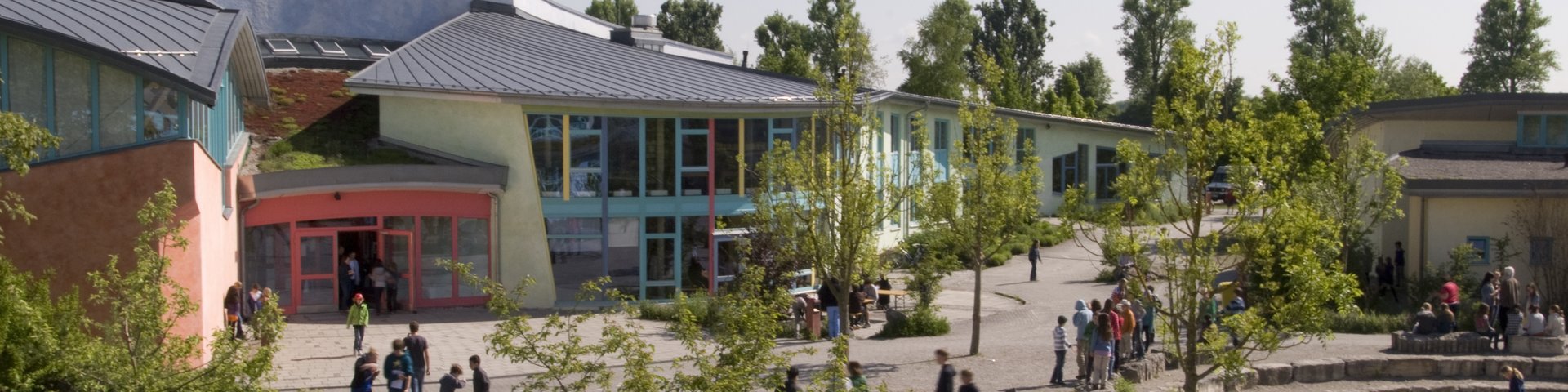 Ein Schulhof mit Gruppen von Schülern, die sich an einem sonnigen Tag vor modernen, farbenfrohen Gebäuden inmitten von Bäumen und Grün versammeln.