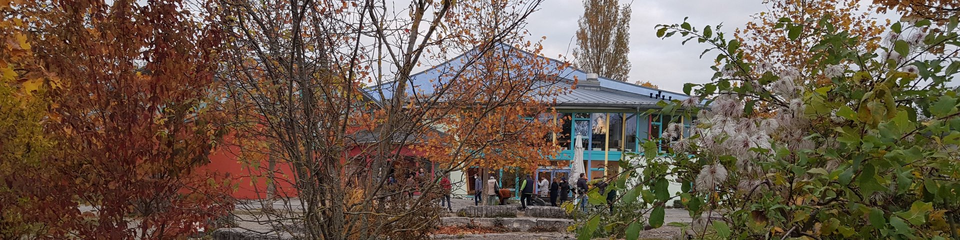 Ein kleines Gebäude mit einem blauen Dach wird teilweise von herbstlichen Bäumen und Büschen verdeckt. Davor bilden große graue Steinblöcke eine niedrige Mauer. Der Himmel ist bedeckt.