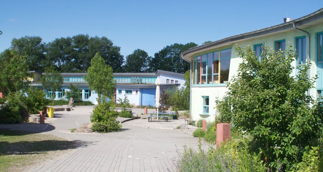 Geschwungene, pastellfarbene Gebäude umgeben einen begrünten Innenhof mit Wegen, Sträuchern, Bänken und Bäumen unter einem klaren blauen Himmel.