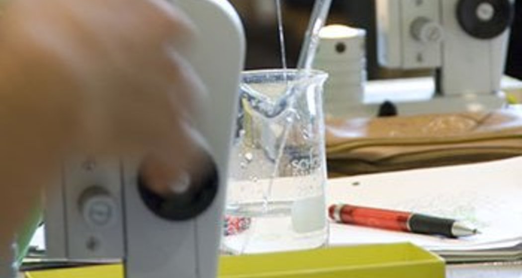 Eine Person benutzt eine Pipette, um Flüssigkeit in ein Becherglas in einem Labor zu transferieren, während Mikroskope und wissenschaftliche Werkzeuge auf dem Tisch stehen und andere Personen im Hintergrund arbeiten.