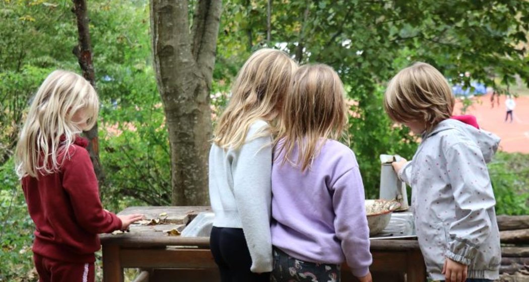 Vier kleine Kinder mit blondem Haar spielen gemeinsam an einem Holztisch im Freien, umgeben von Bäumen und herabgefallenen Blättern. Sie scheinen einer Beschäftigung nachzugehen, möglicherweise mit Gegenständen aus der Natur.