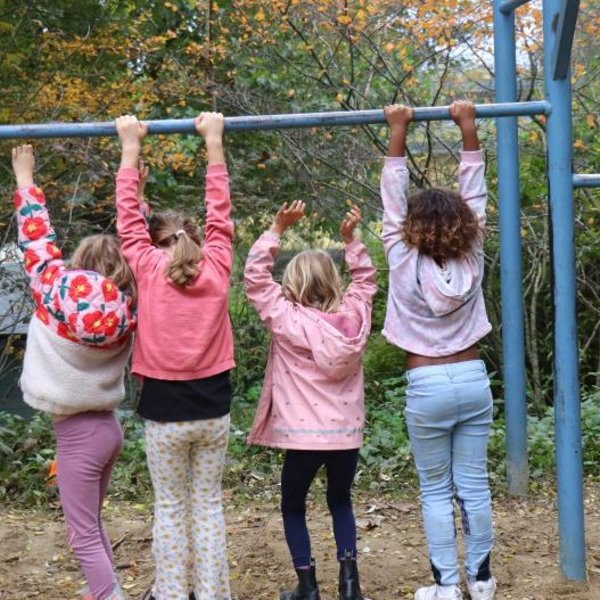 Sechs Kinder hängen im Freien an einer horizontalen Stange, von der Kamera abgewandt. Fünf halten sich mit den Händen fest, während eines, ganz rechts, kopfüber an den Knien hängt. Der Hintergrund ist mit Bäumen und Laub bedeckt.