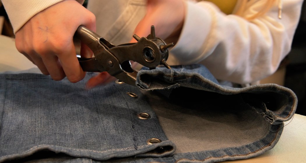 Eine Person verwendet einen rotierenden Lederlocher, um Löcher in ein Stück blauen Jeansstoff zu stanzen, der am Rand bereits mit Metallösen versehen ist. Die Person trägt ein helles Sweatshirt.