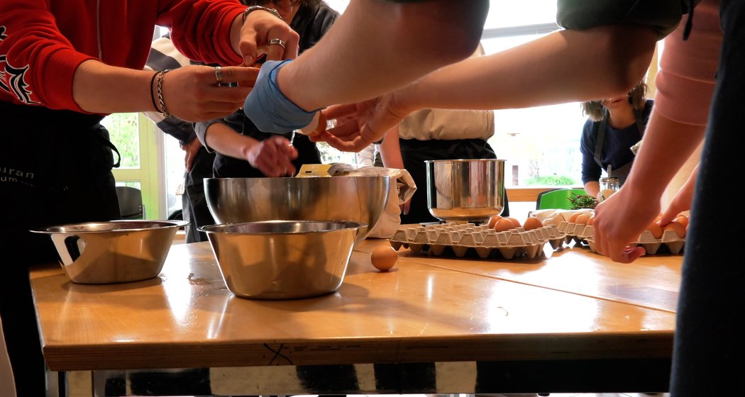 Mehrere Personen stehen um einen Holztisch herum, auf dem Rührschüsseln, Eier und Küchenutensilien verstreut sind, und bereiten das Essen vor. Einige Hände schlagen Eier auf, während andere dabei helfen oder Zutaten mischen.