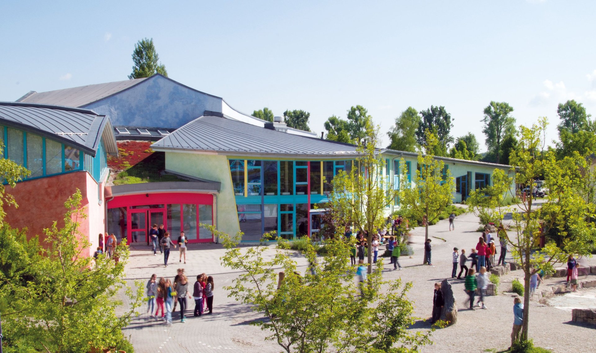 Ein farbenfrohes Schulgebäude mit großen Fenstern und Bäumen rundherum, mit Gruppen von Kindern und Erwachsenen, die an einem sonnigen Tag draußen spazieren gehen und sich versammeln.
