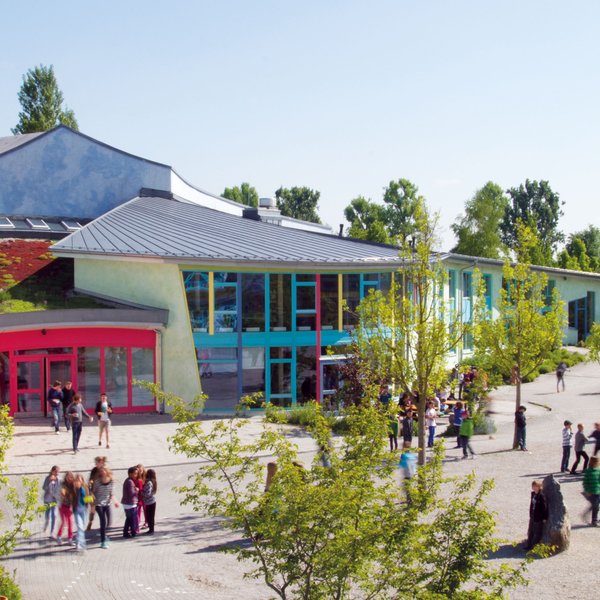 Ein farbenfrohes Schulgebäude mit großen Fenstern und Bäumen rundherum, mit Gruppen von Kindern und Erwachsenen, die an einem sonnigen Tag draußen spazieren gehen und sich versammeln.