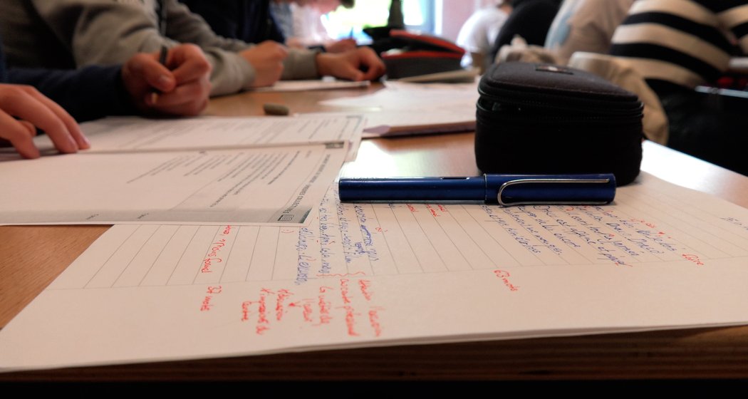 Nahaufnahme eines Schreibtisches mit handschriftlichen Notizen, einem blauen Stift und einem Federmäppchen, während mehrere Schüler im Hintergrund in einem Klassenzimmer an Papieren schreiben.