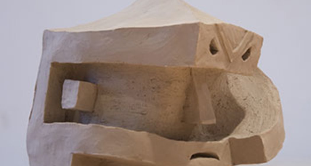 Abstrakte Tonskulptur mit geometrischen Formen, darunter rechteckige Ausschnitte und eine schräge Spitze, die an ein stilisiertes menschliches Gesicht mit kantigem Kiefer und ausgeprägter Stirn erinnert. Das Kunstwerk steht auf einer glatten, hellen Oberfläche.