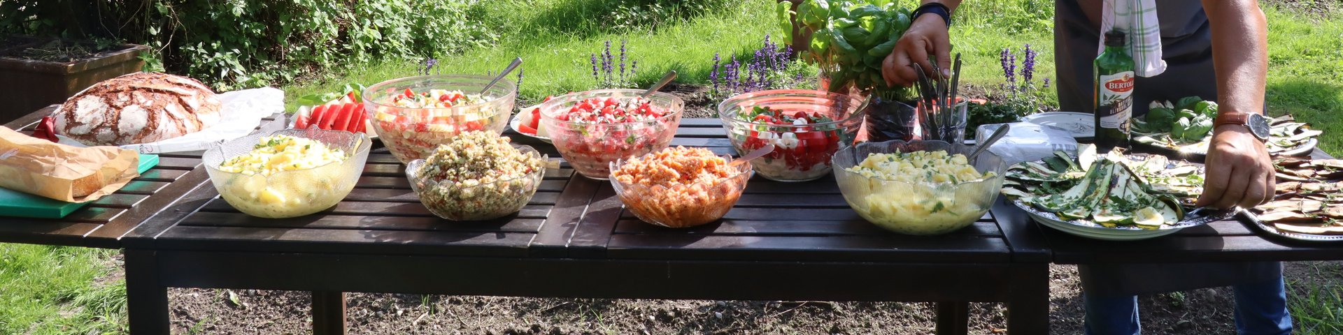 Ein Mann arrangiert Schüsseln mit Salaten und Gerichten auf einem Tisch im Freien, der für eine Mahlzeit in einem Garten gedeckt ist, mit Bäumen und einem rustikalen Schuppen im Hintergrund. Ein Handtuch ist über seine Schulter drapiert.