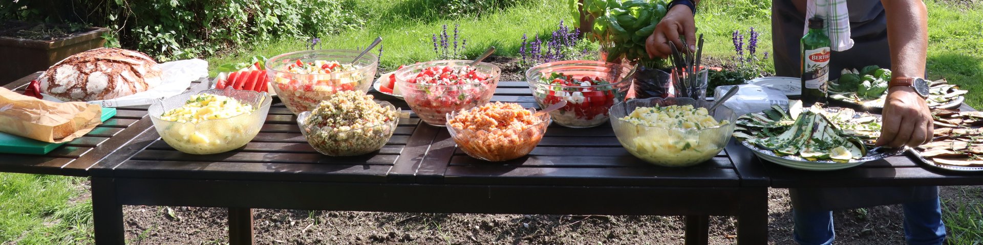 Ein Mann arrangiert Schüsseln mit Salaten und Gerichten auf einem Tisch im Freien, der für eine Mahlzeit in einem Garten gedeckt ist, mit Bäumen und einem rustikalen Schuppen im Hintergrund. Ein Handtuch ist über seine Schulter drapiert.