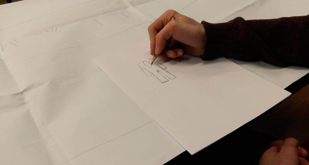 Eine Person, die einen dunklen Pullover trägt, zeichnet mit einem Bleistift einen einfachen Umriss eines Gebäudes auf ein weißes Blatt Papier. Auf dem Tisch sind mehrere Blätter mit schwachen Skizzen ausgebreitet.