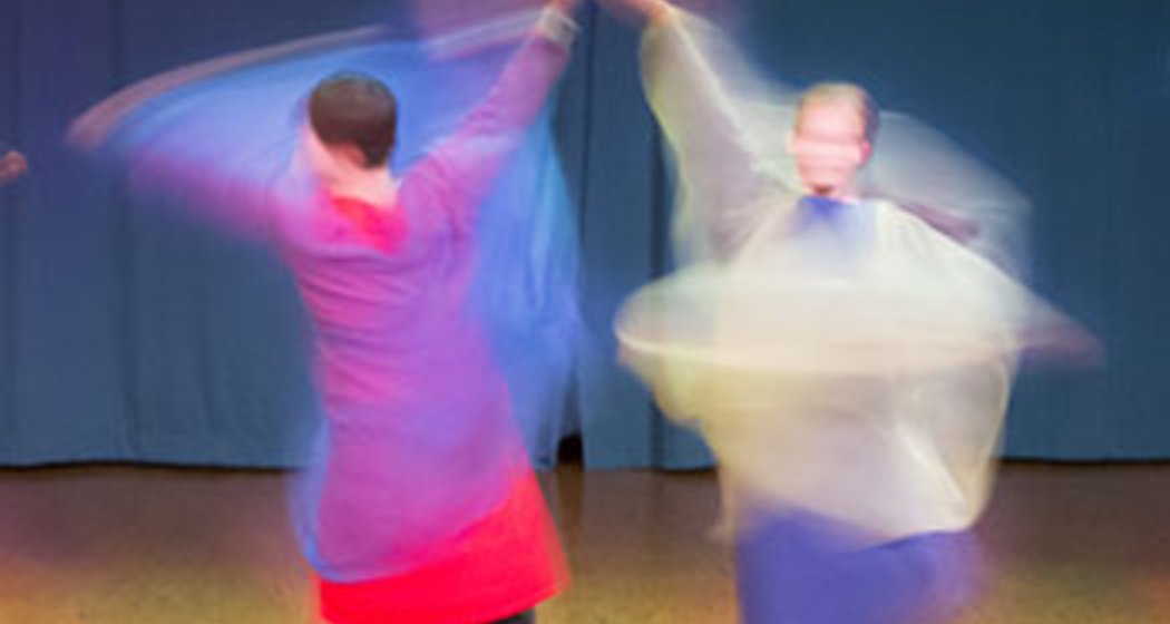 Zwei Personen in farbenfroher traditioneller Kleidung wirbeln und tanzen auf einer Bühne mit unscharfen Bewegungen, wobei sie mit ihren Kleidern fließende, durchscheinende Formen erzeugen. Im Hintergrund ist ein schlichter blauer Vorhang zu sehen.