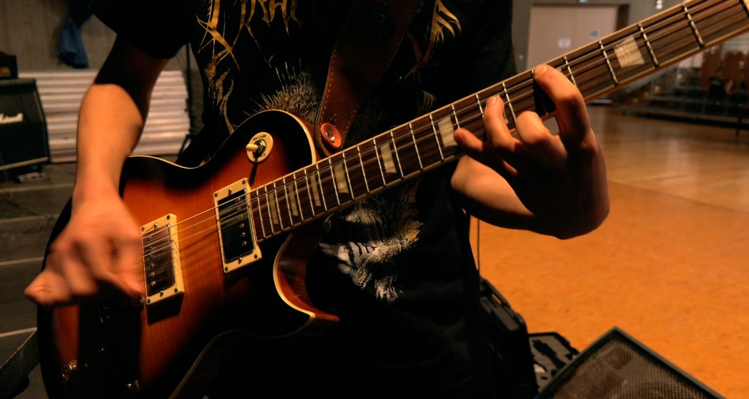 Eine Person, die ein dunkles grafisches T-Shirt trägt, spielt eine E-Gitarre und konzentriert sich dabei auf ihre linke Hand, die die Saiten auf dem Griffbrett anschlägt. Der Hintergrund ist ein Innenraum und leicht unscharf.