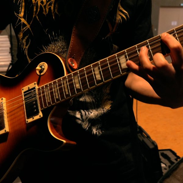 Eine Person, die ein dunkles grafisches T-Shirt trägt, spielt eine E-Gitarre und konzentriert sich dabei auf ihre linke Hand, die die Saiten auf dem Griffbrett anschlägt. Der Hintergrund ist ein Innenraum und leicht unscharf.