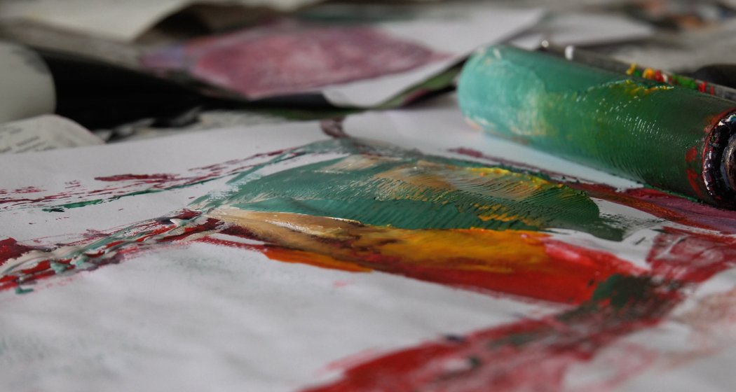 Eine Nahaufnahme eines Farbrollers, der grüne, gelbe und rote Farbe auf Papier aufträgt, wobei andere Kunstgegenstände und Kunstwerke im Hintergrund verschwimmen.