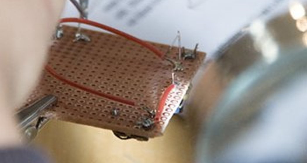 Eine Person arbeitet mit einem Lötkolben an einer elektronischen Leiterplatte mit roten Drähten, während sie die Platte mit einem Hilfswerkzeug festhält. Ein unscharfes Dokument und Werkzeuge sind im Hintergrund zu sehen.
