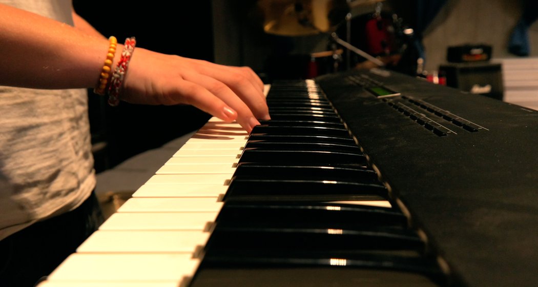 Nahaufnahme einer Person, die ein schwarz-weißes Keyboard spielt, möglicherweise ein elektrisches Klavier oder einen Synthesizer, mit einem Schlagzeug im Hintergrund. Die Person trägt bunte Perlenarmbänder.