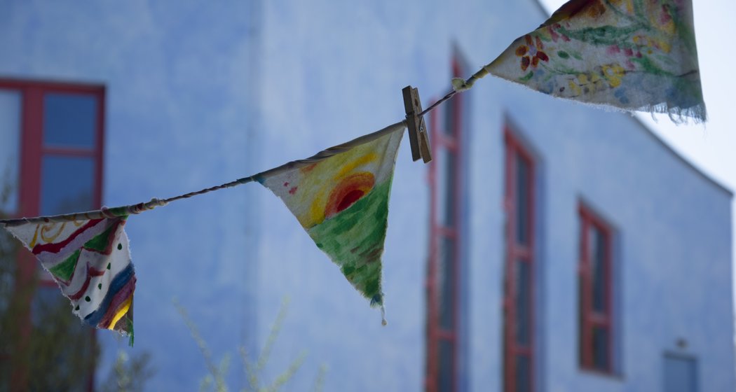 Bunte, handbemalte Stoffwimpel hängen mit einer Wäscheklammer an einer Schnur, vor einem verschwommenen blauen Gebäude mit roten Fensterrahmen im Hintergrund.