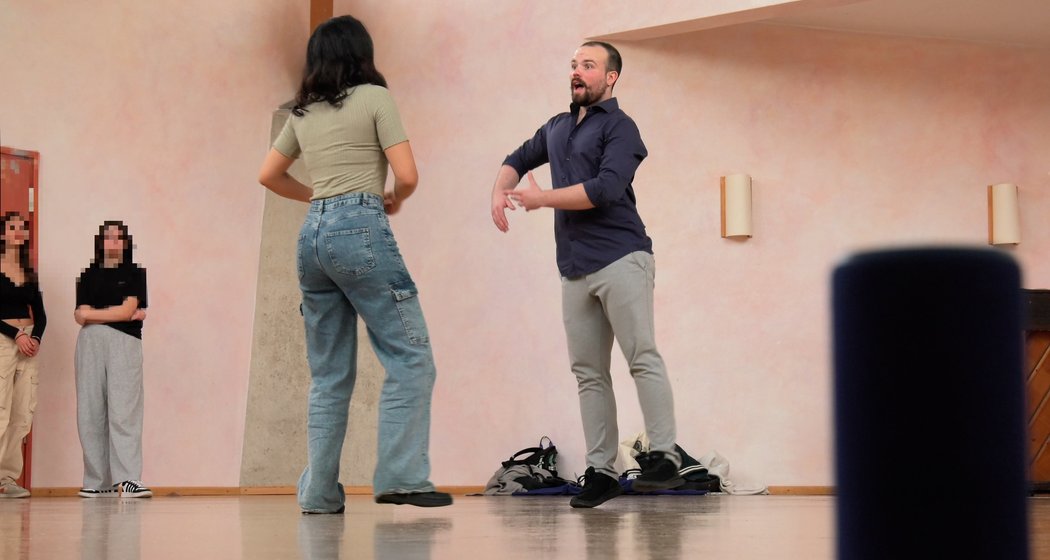 Zwei Personen tanzen zusammen in einem Studio mit Holzboden, während zwei weitere Personen im Hintergrund zusehen. Kleidung und Schuhe liegen in der Nähe auf dem Boden. Der Raum hat hellrosa Wände und ist minimalistisch eingerichtet.
