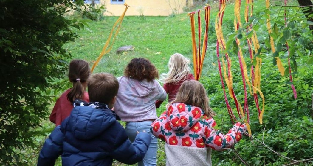 Fünf Kinder rennen im Freien auf ein gelbes Gebäude zu und laufen dabei durch bunte Bänder, die von den Ästen der Bäume herabhängen. Die Szene ist üppig und grün und suggeriert eine spielerische, abenteuerliche Atmosphäre in der Natur.